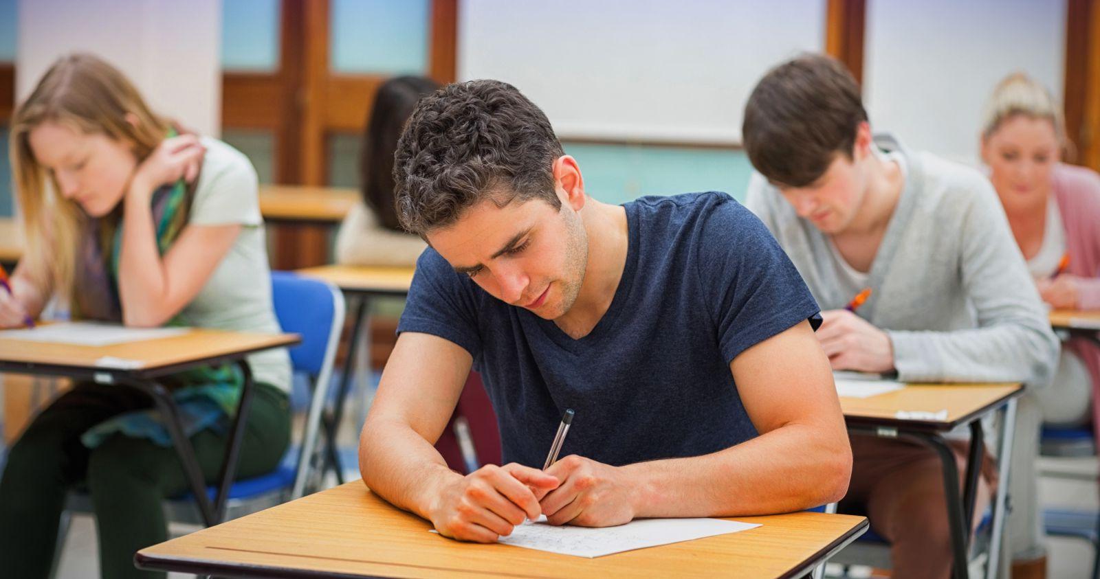 Two male and two female Caucasian students wearing casual clothing sit at desks in a classroom while writing on paper like they are taking a test.