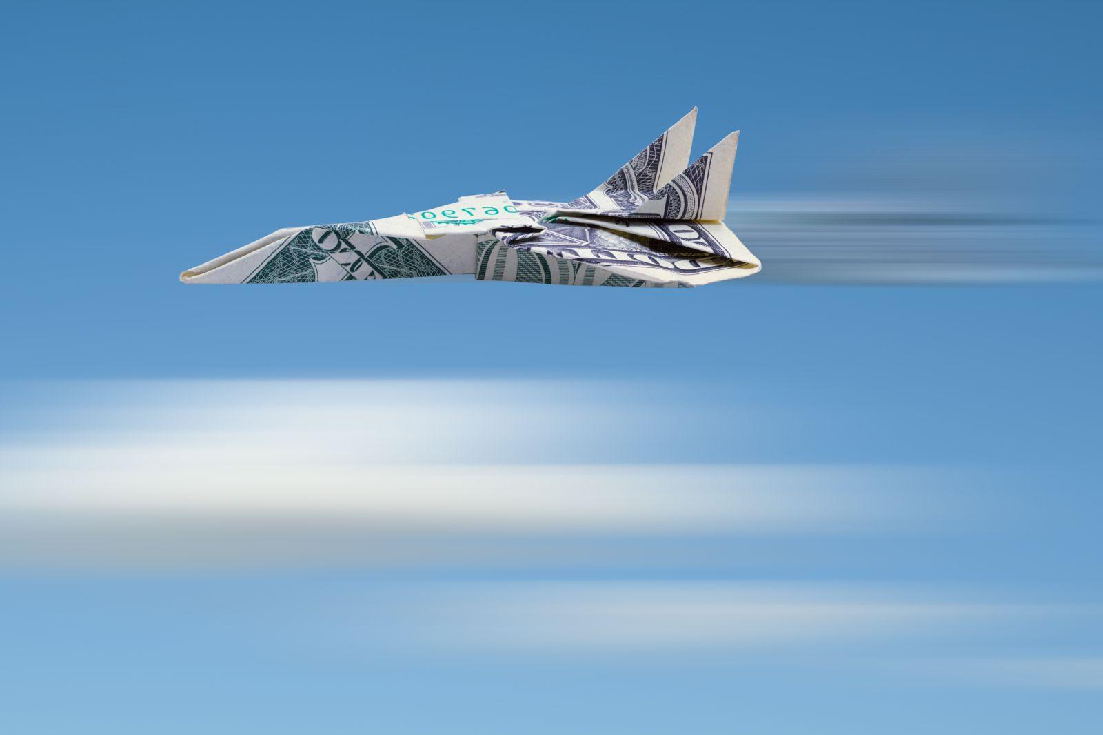 A paper airplane made from a 1-dollar bill is set against a sky-blue backdrop with white blur lines below that look like blurred clouds and black and white blur lines behind the engines to represent the plane flying at great speed.