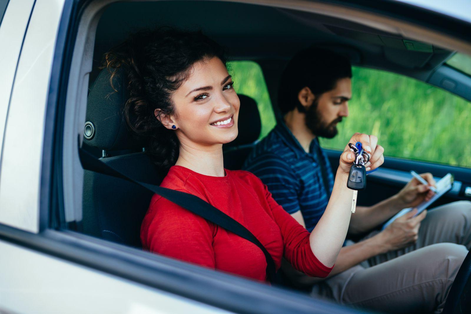 A smiling Caucasian female with black hair wearing a red long-sleeved t-shirt sits in a silver car holding up keys in her right hand while a bearded Caucasian male also with black hair and wearing business casual clothing writes on a paper using a clipboard.